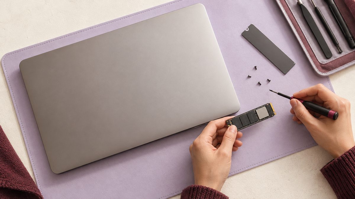 Hands inspect a storage module beside a laptop and precision tools.